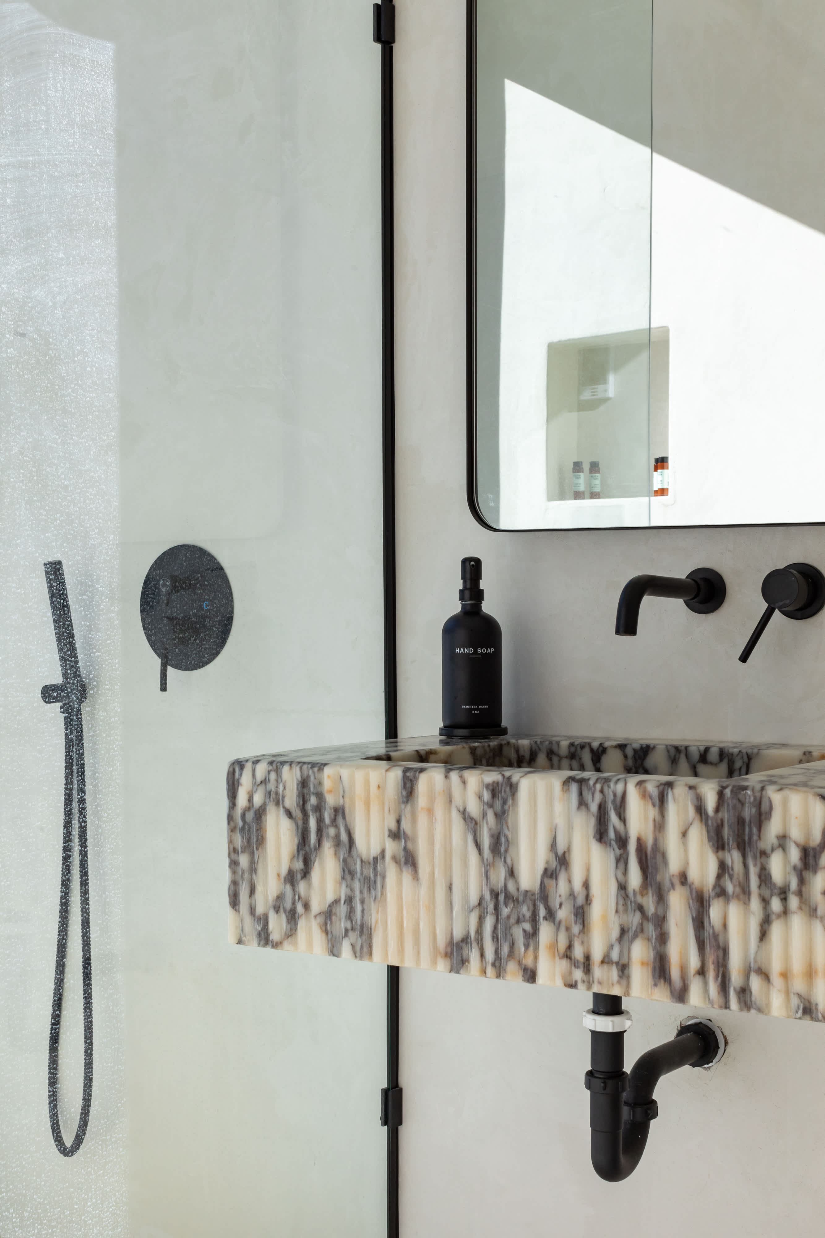 A modern bathroom features a unique fluted marble vanity with a black wall-mounted faucet and a black soap dispenser. A black-framed mirror hangs above, and a black shower head and handheld sprayer are visible in the adjacent glass-enclosed shower.