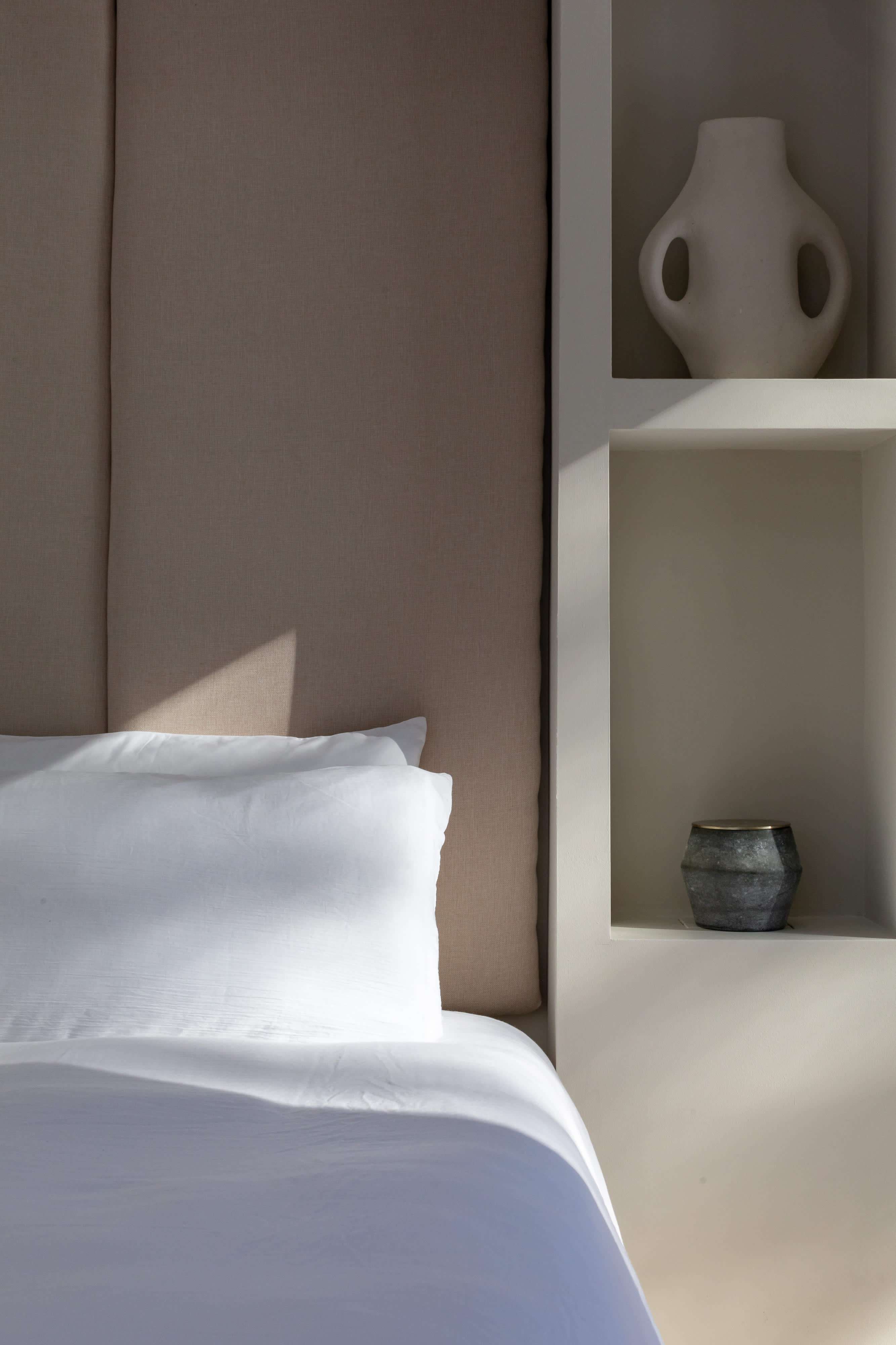 A close-up of the bed with crisp white linens and a beige upholstered headboard, adjacent to a built-in shelf displaying a white ceramic vase and a dark textured candle holder, bathed in natural light.