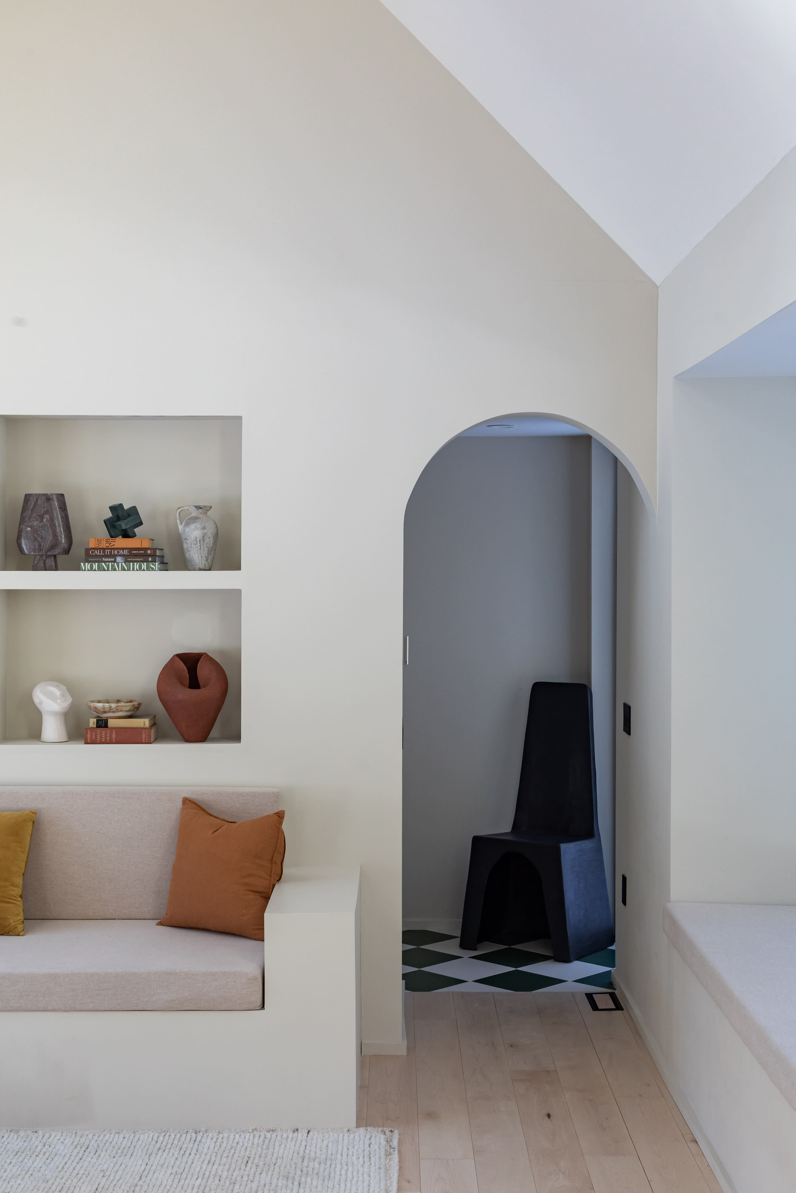 A minimalist living area with light cream walls, featuring a built-in sofa with neutral and rust-colored cushions, and recessed shelving displaying decorative vases and books. An arched doorway leads to another room with a black chair and checkered floor.