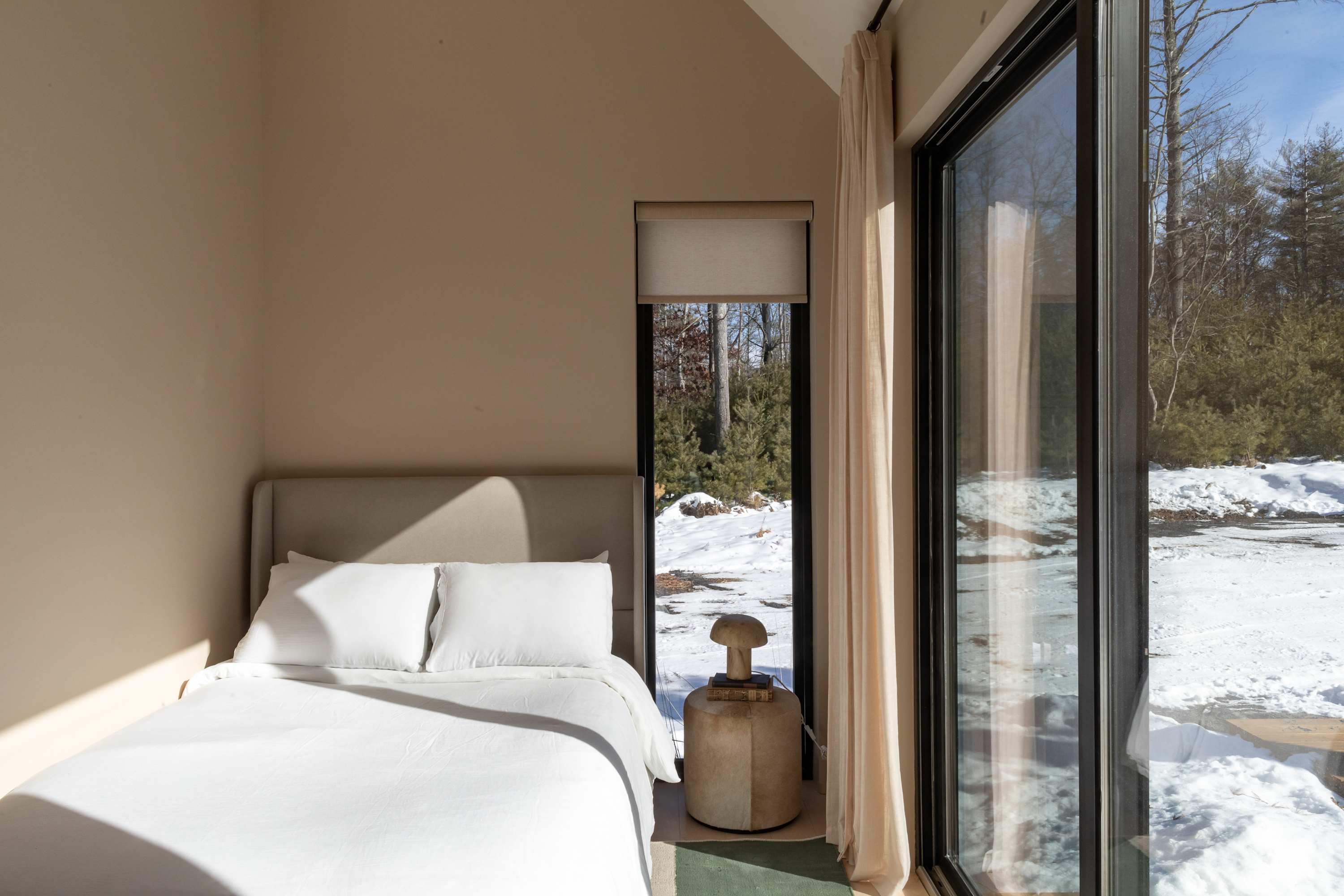 A bright bedroom with light-colored walls features a bed with white linens and a gray headboard, next to a tall window and a glass door offering views of a snowy, wooded exterior. A round nightstand with a unique lamp sits between the bed and the window.