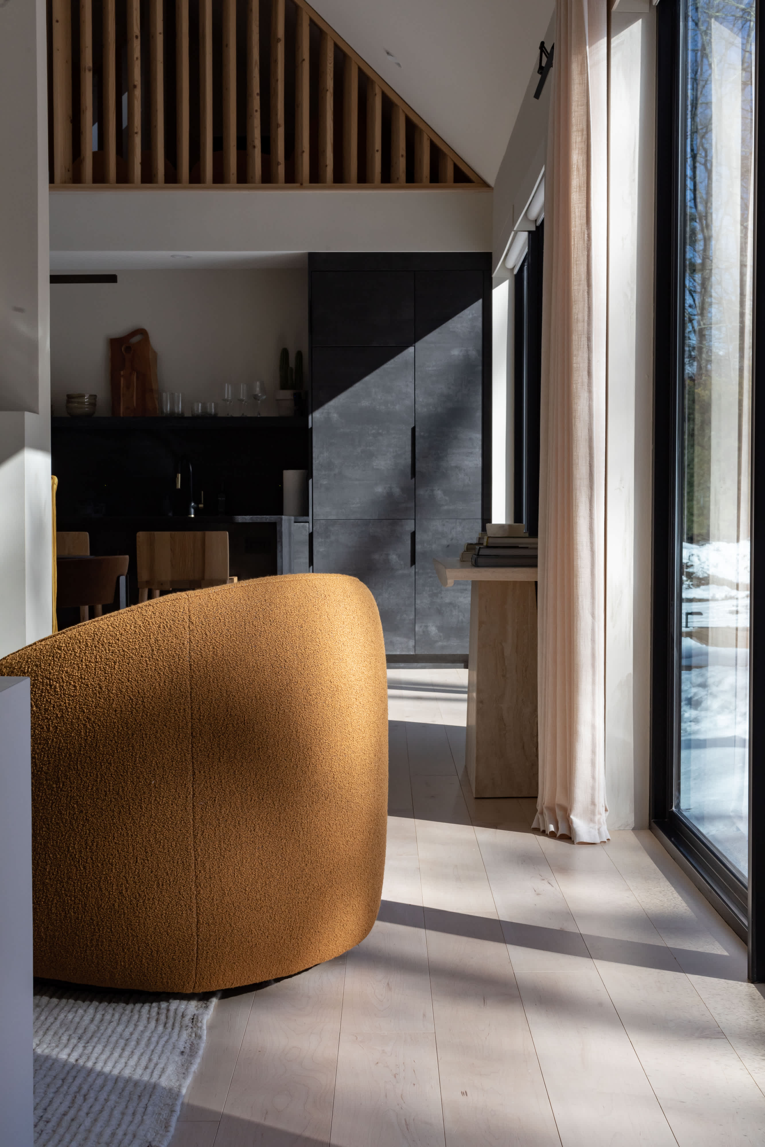 A modern living space with light wood floors, a curved mustard yellow armchair, and a view into a dark, minimalist kitchen with a slatted wood ceiling detail. Sunlight streams through a large window, highlighting the clean lines and contemporary design.