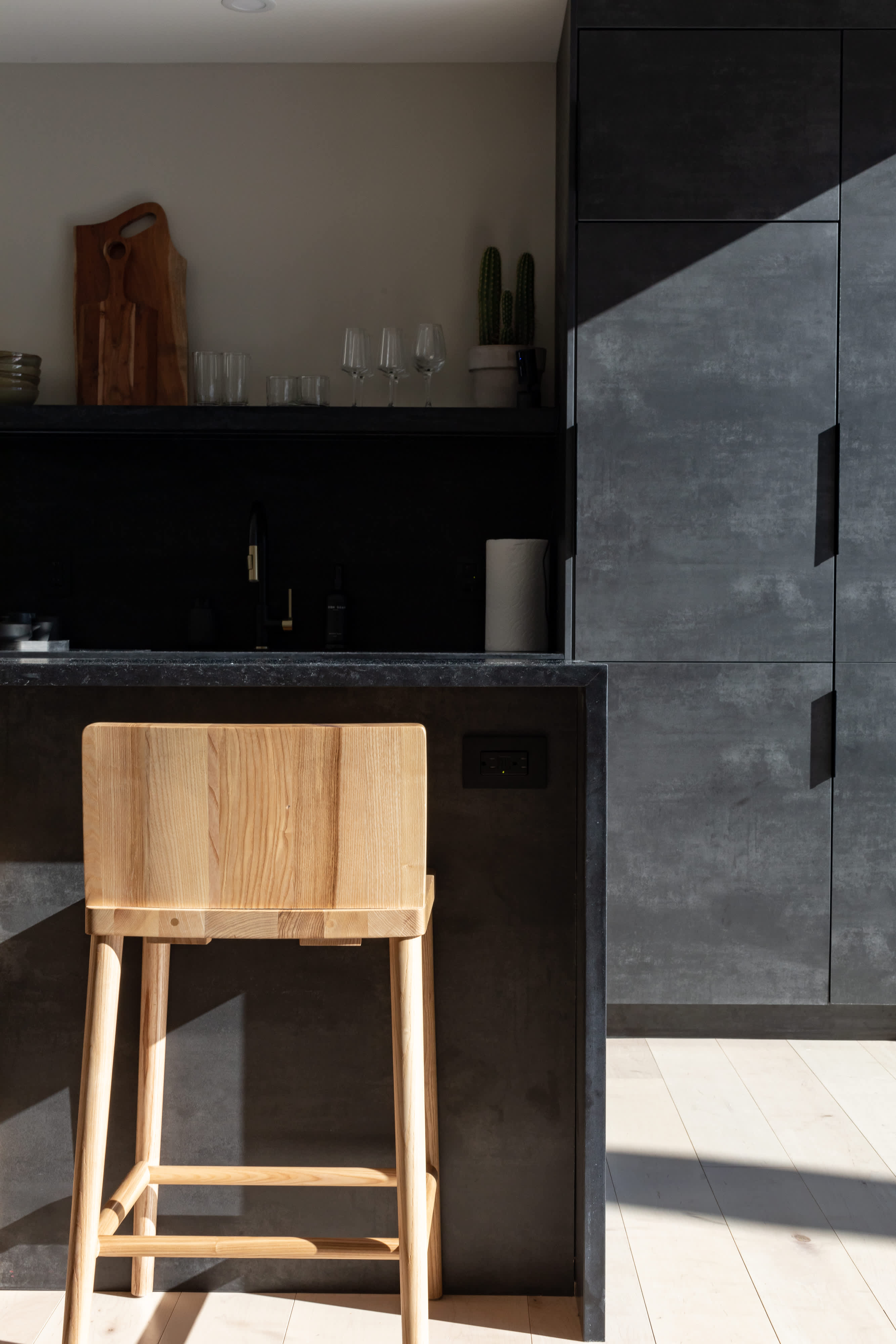 A close-up of a minimalist kitchen island with dark countertops and cabinetry, featuring a light wooden bar stool. Sunlight casts strong shadows across the textured dark surfaces, highlighting the modern design.