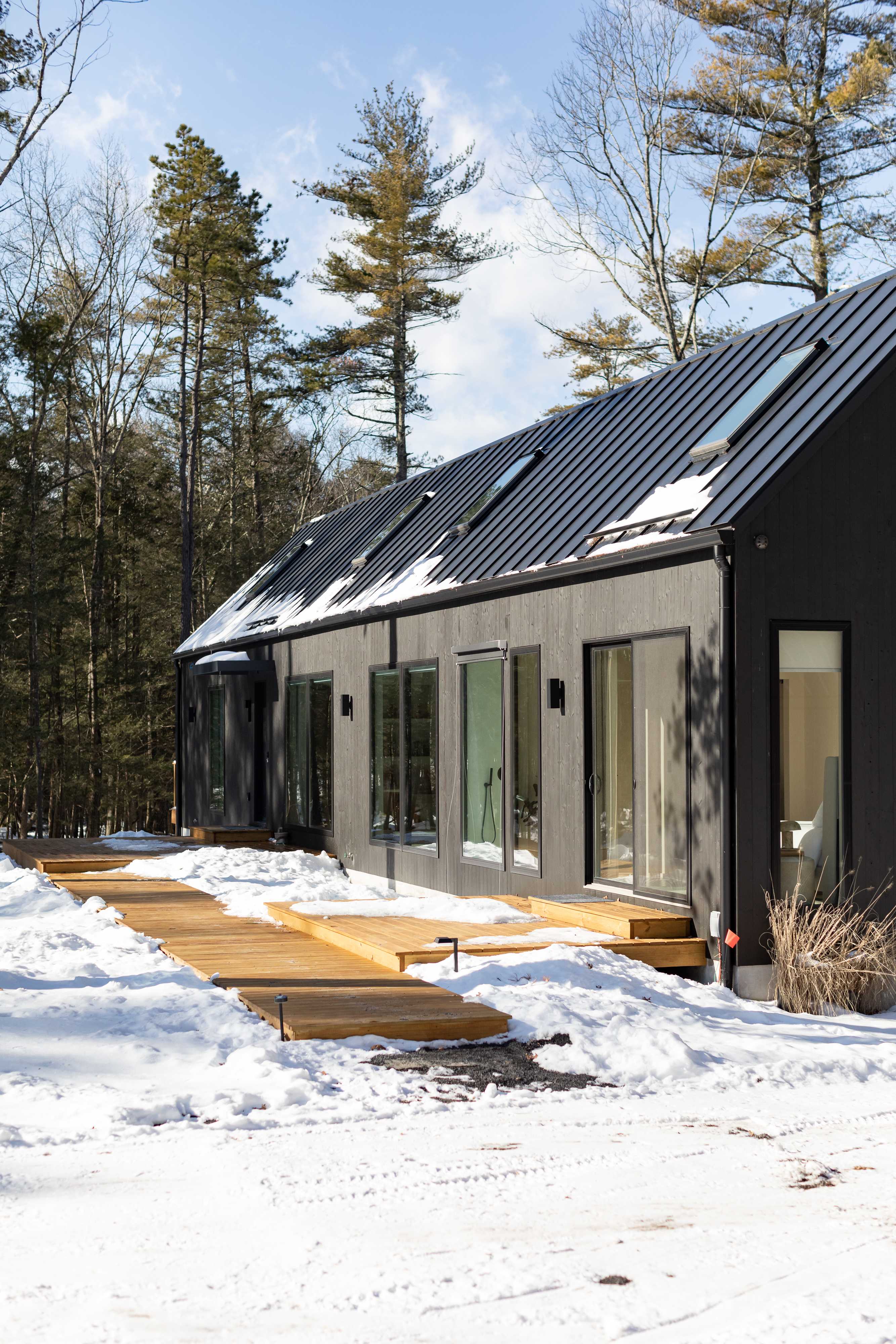 A modern, dark-sided house with a black metal roof and multiple skylights, featuring a large wooden deck partially covered in snow. Tall pine trees and bare deciduous trees surround the property.