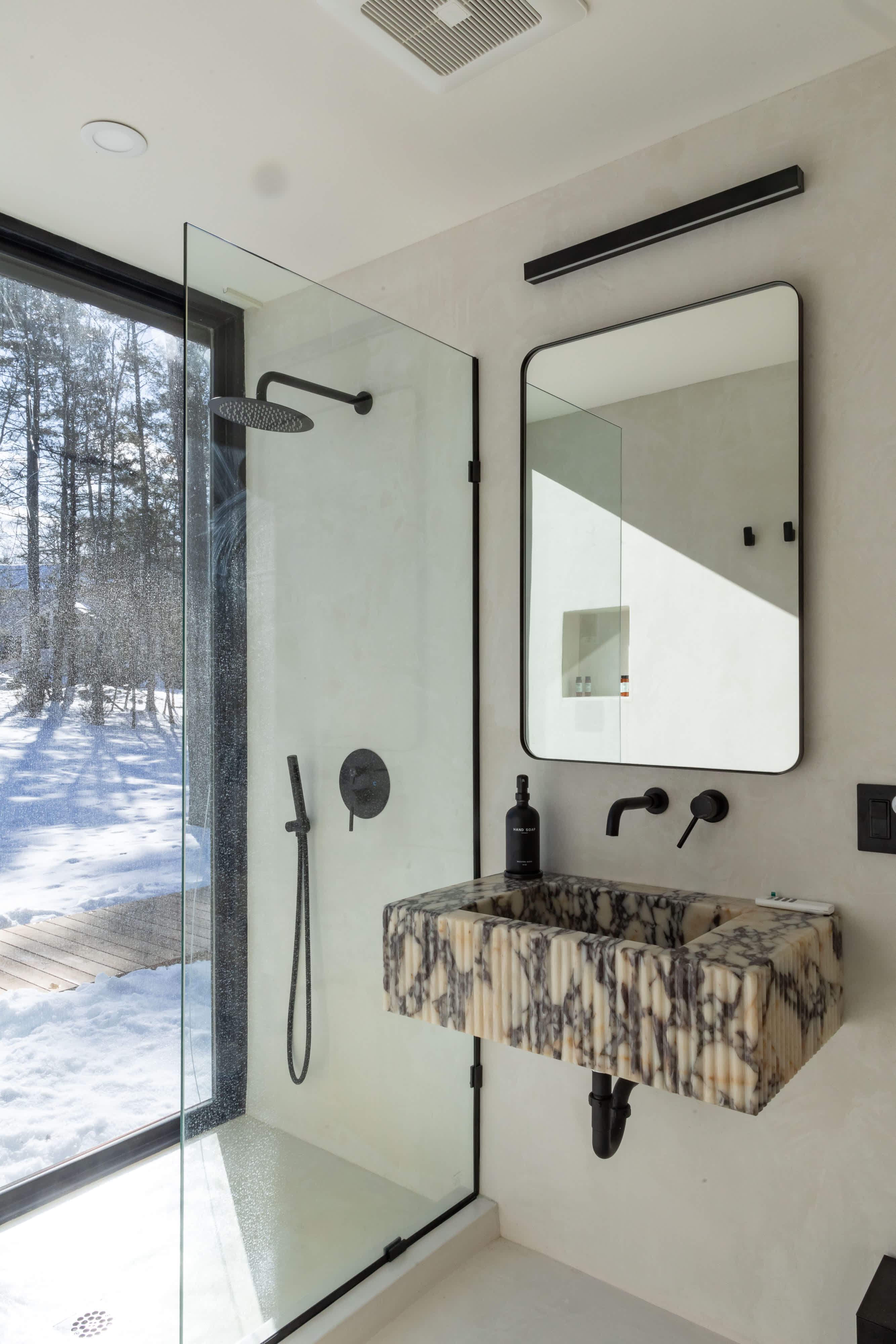A contemporary bathroom features a unique fluted marble vanity with black fixtures and a black-framed mirror. A spacious glass-enclosed shower includes a large window offering a view of a snowy, wooded landscape, creating an immersive experience.