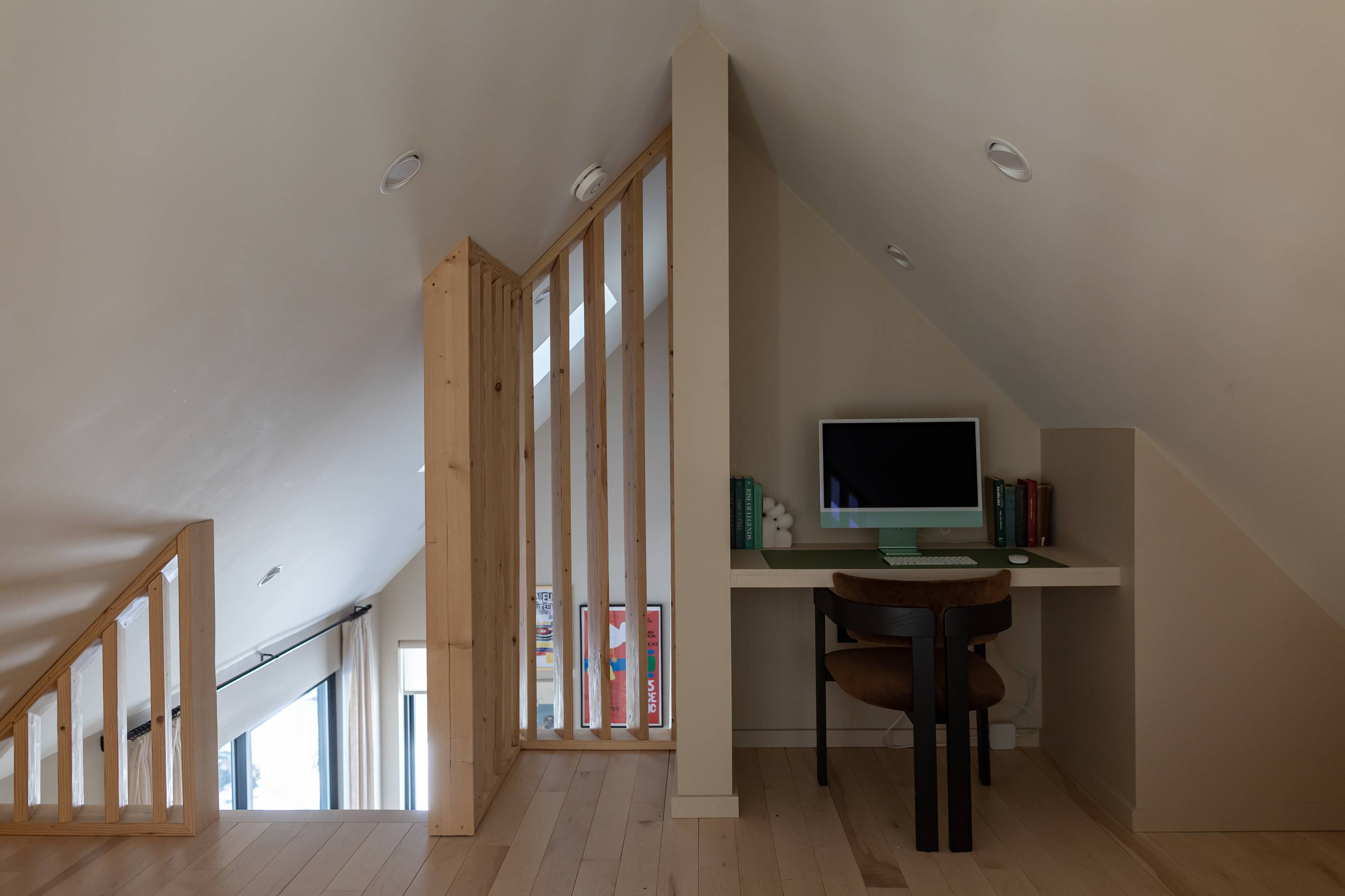 A bright attic office space features a built-in desk with a light green iMac and various books, complemented by a dark brown chair. A wooden slat partition separates the workspace from another area, all under a sloped ceiling with recessed lighting.