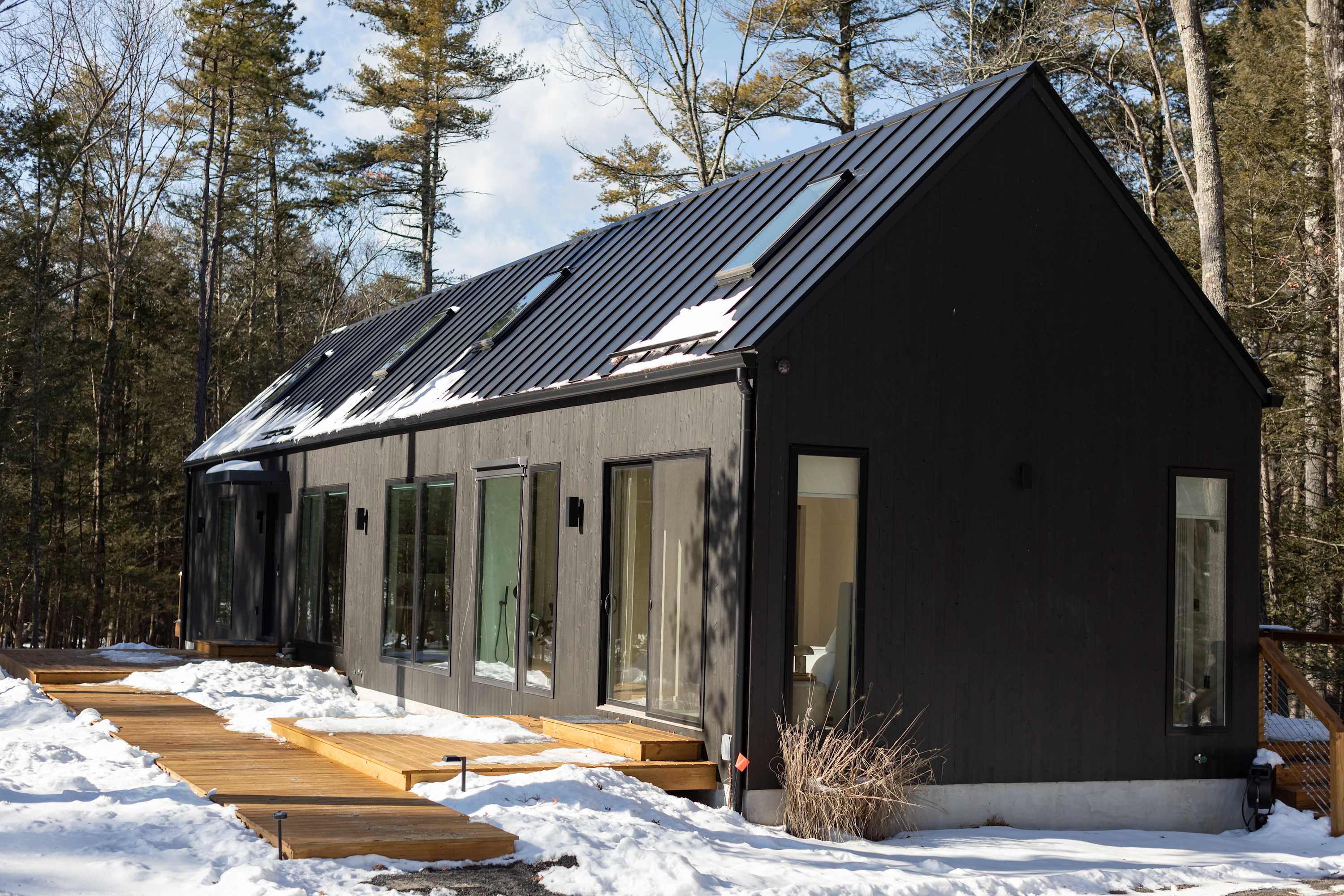 A long, modern black house with a pitched black metal roof and several skylights, surrounded by a snowy forest. A multi-level wooden deck extends along the side of the house, partially covered in snow.