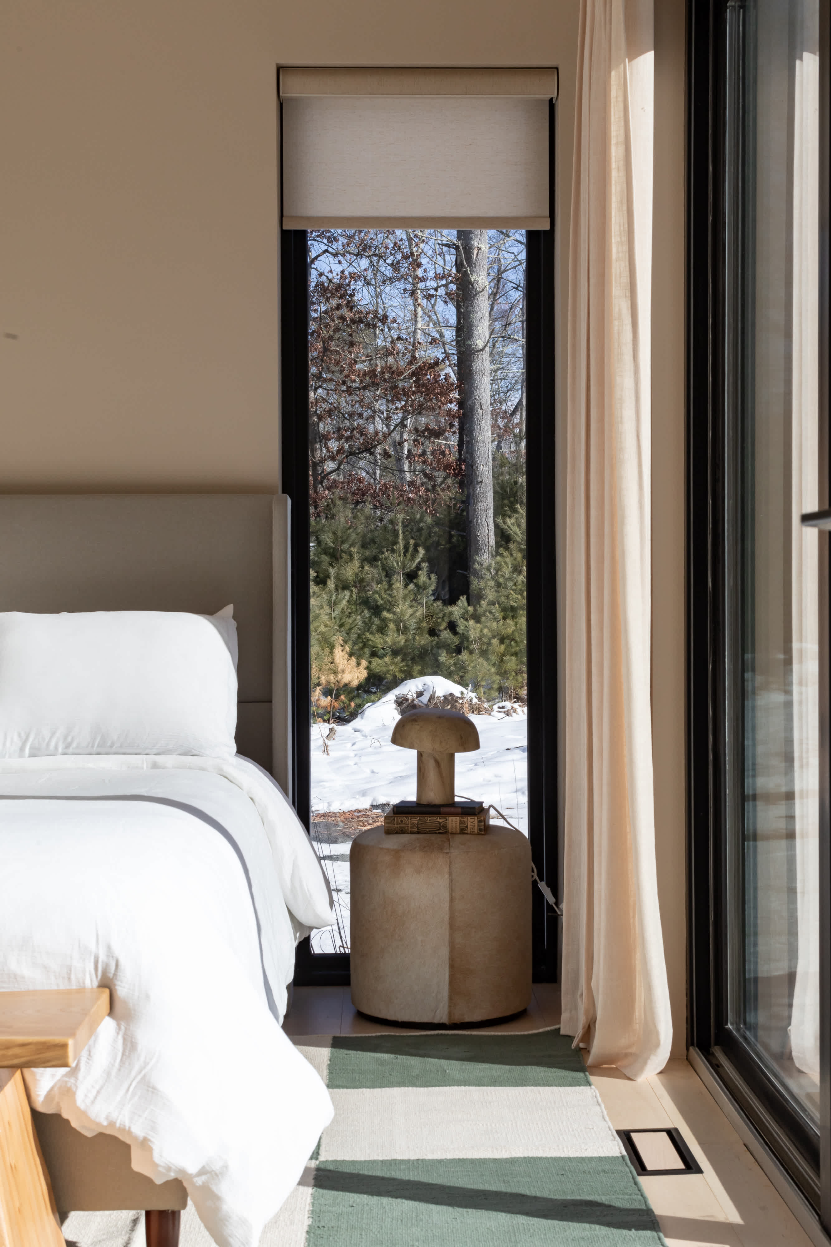A serene bedroom features a bed with crisp white linens and a light gray headboard, positioned next to a tall window overlooking a snowy, wooded landscape. A unique mushroom-shaped lamp sits on a round, textured nightstand beside the bed.