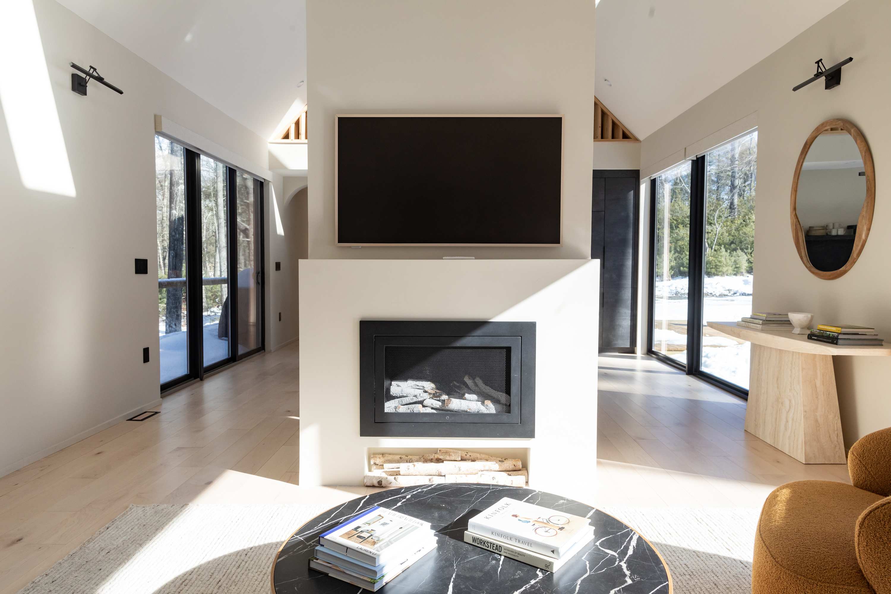 A bright living room centered around a modern fireplace with a television mounted above, flanked by large windows offering views of the snowy outdoors. Light wood floors and a minimalist aesthetic define the space.