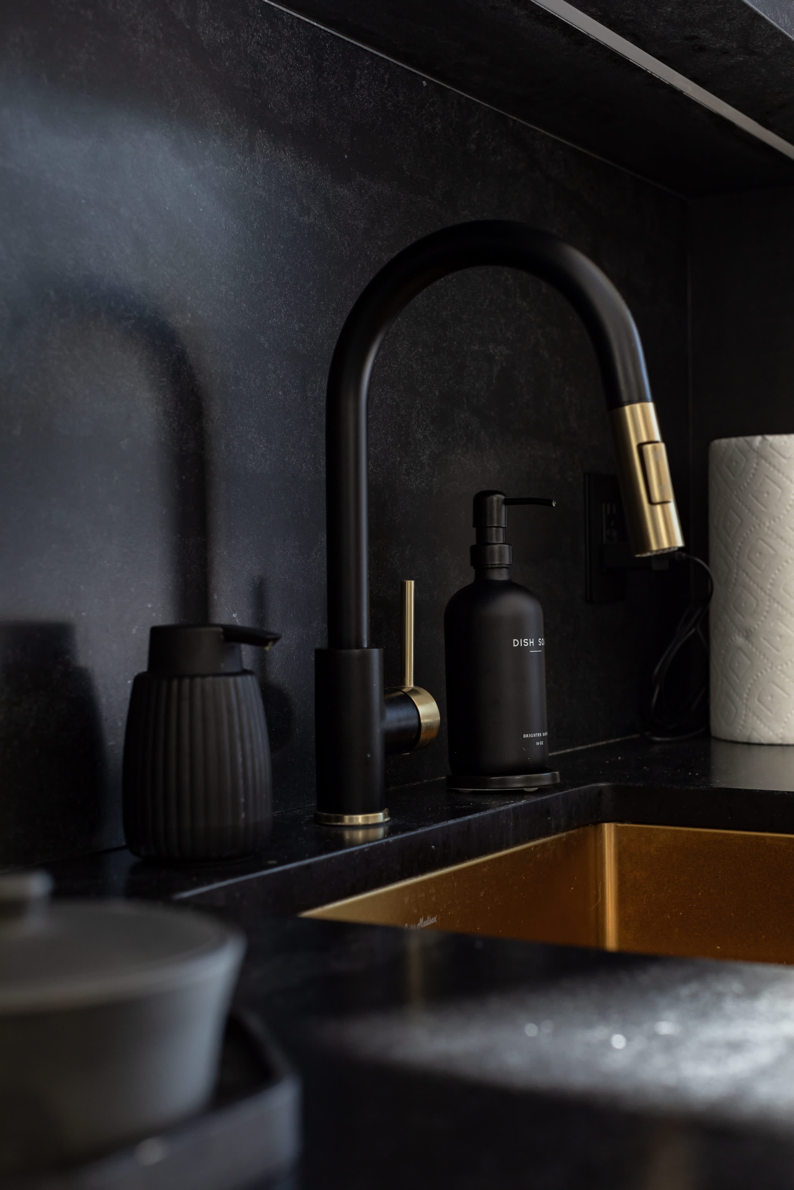 A close-up of a modern kitchen sink area with a black gooseneck faucet featuring gold accents, set against a dark countertop and backsplash. A gold sink basin and various kitchen accessories are visible.