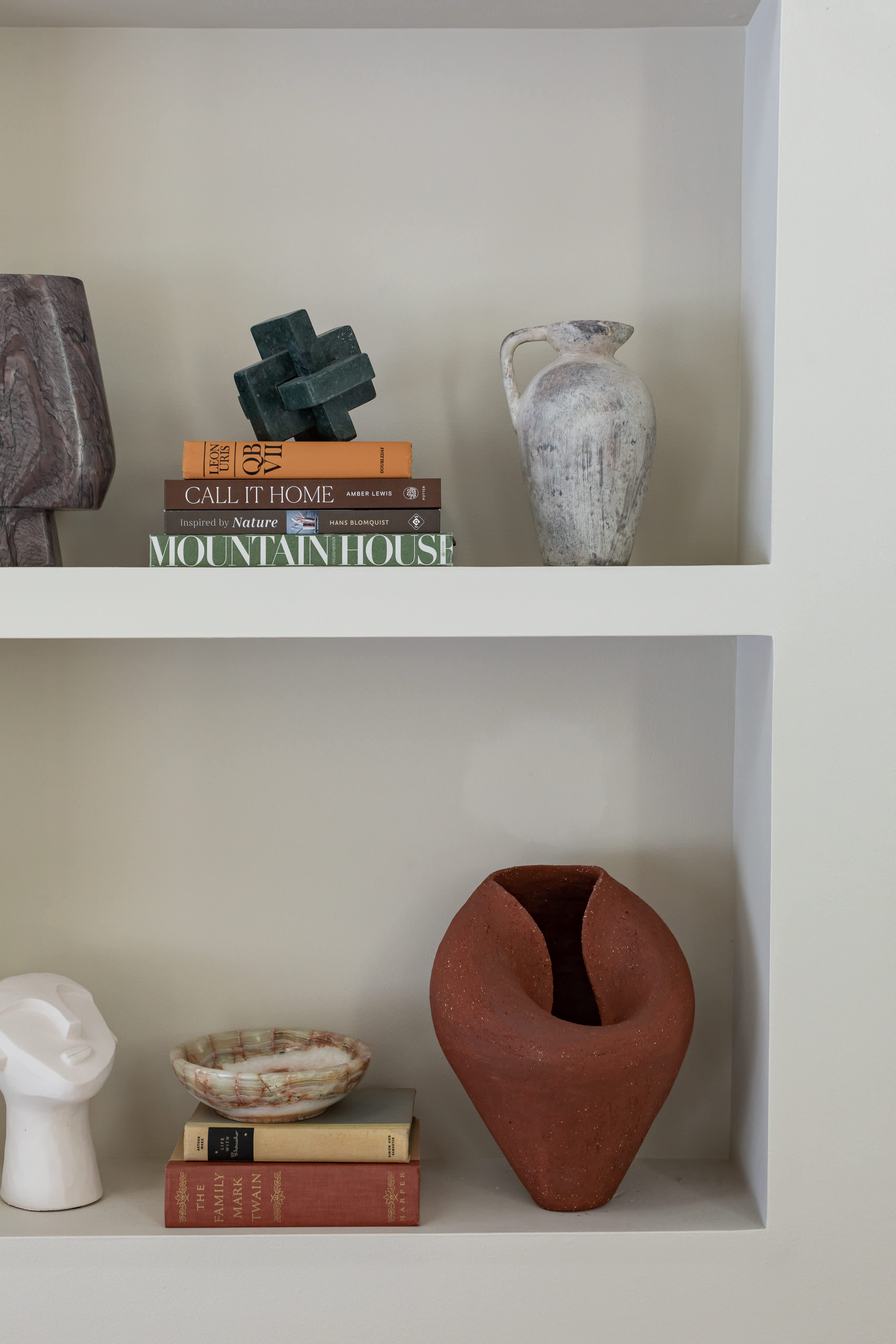 Built-in shelving displays an eclectic collection of decorative objects, including abstract sculptures, a rustic vase, and various art and design books. The shelves are painted a light neutral color, complementing the diverse textures and colors of the items.