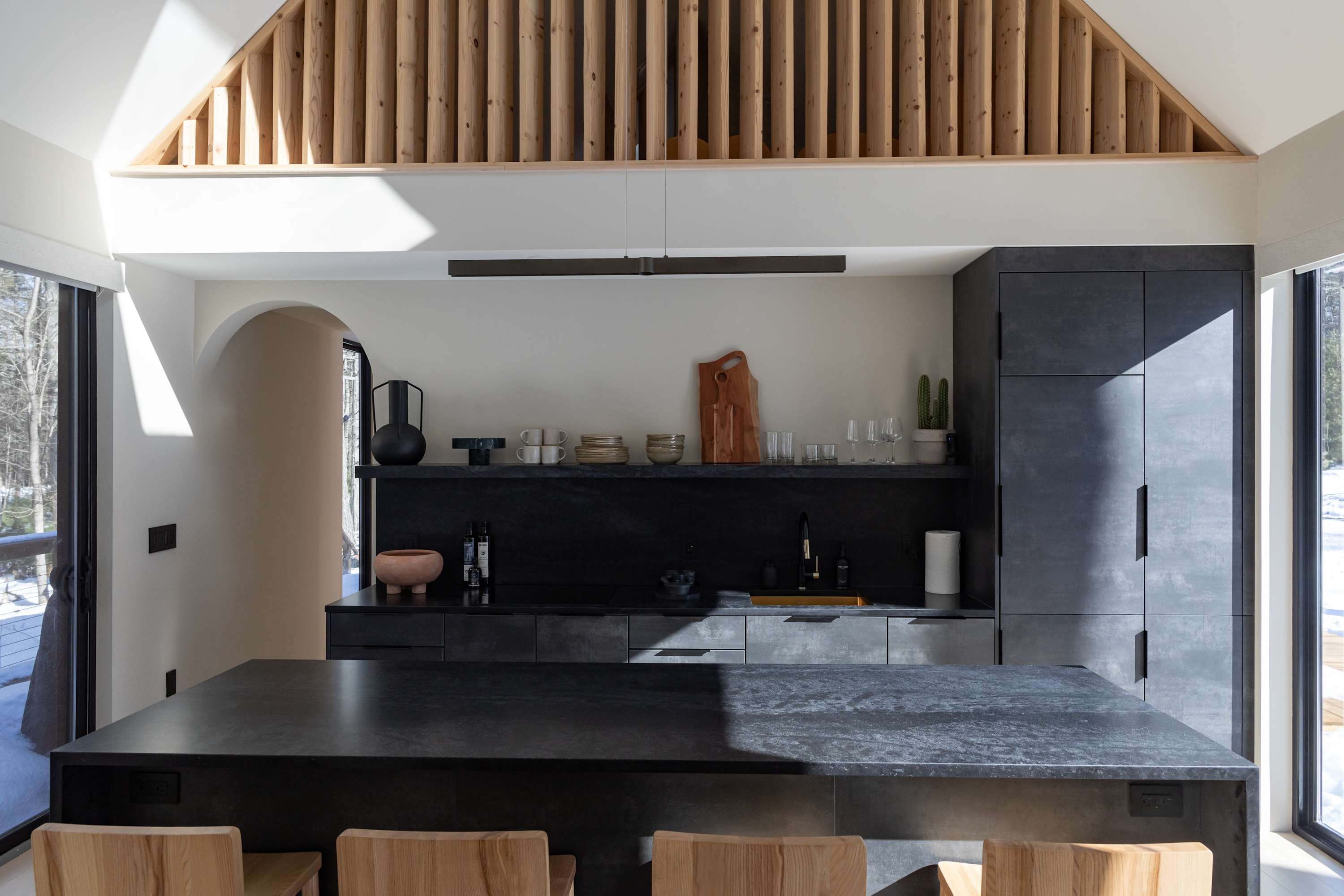 A sleek, modern kitchen with dark, textured cabinetry and countertops, featuring an island with wooden bar stools and an open shelf displaying dishes. A unique slatted wood ceiling detail adds warmth, and an arched doorway leads to another room.