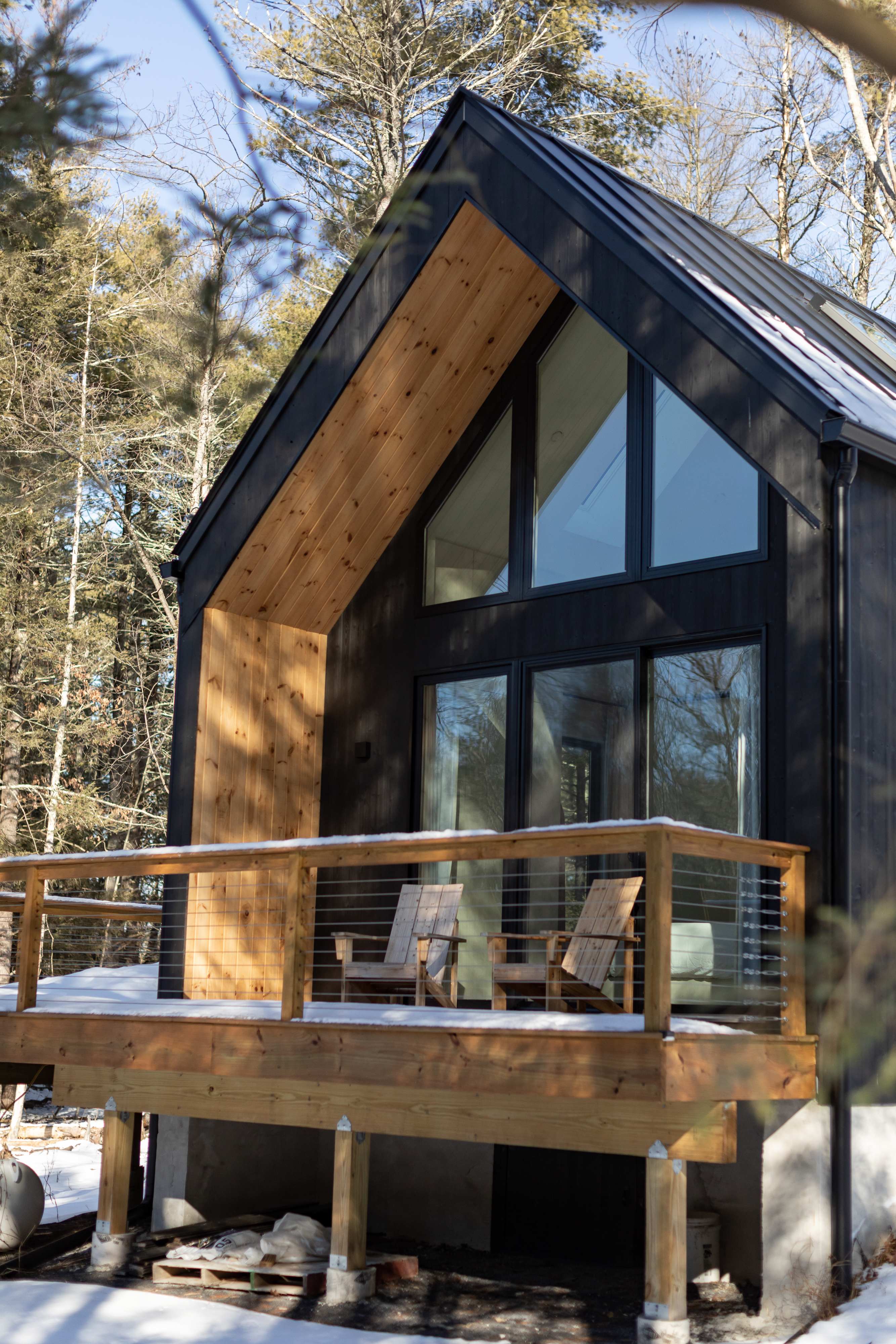 A striking black modern cabin with a steep gabled roof and large windows, featuring a wooden deck with two Adirondack chairs, surrounded by snow-dusted pine trees. The exterior combines dark siding with warm wood accents under the eaves.