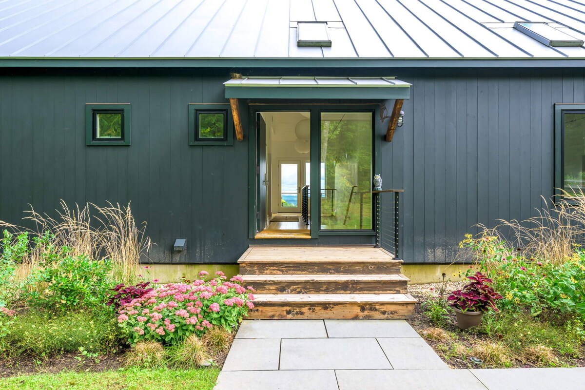 Inviting front entrance of a dark-sided house with wooden steps leading up to a glass door, flanked by landscaped garden beds with pink flowers and tall grasses, under a small metal awning.