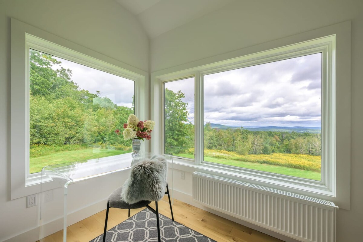 A corner of a bright bedroom with light hardwood floors, featuring a modern chair with a furry throw and a clear acrylic table, positioned by expansive corner windows that offer panoramic views of rolling mountains and a vibrant green landscape.
