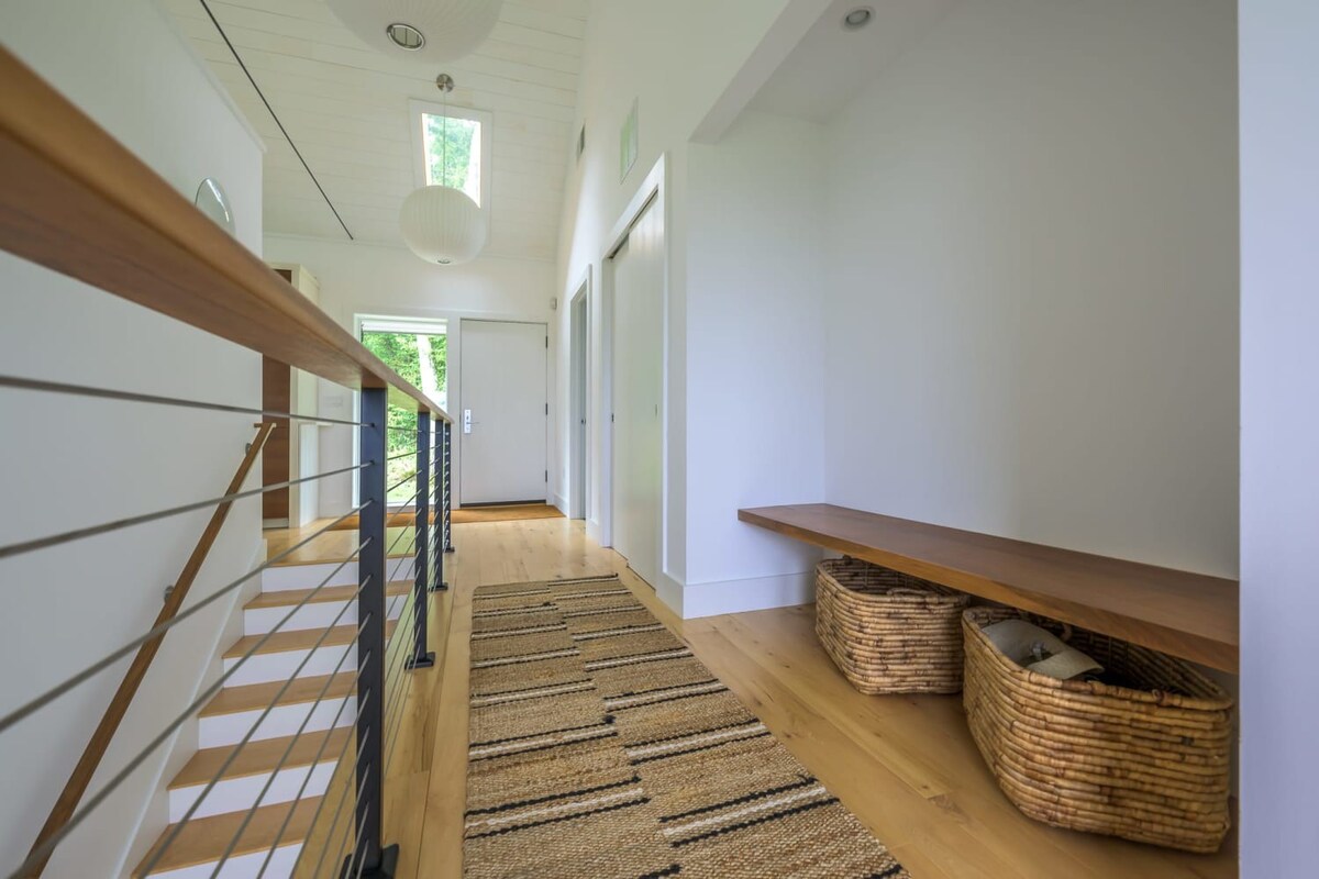 Bright hallway with light wood flooring, a modern staircase featuring cable railings and a wooden handrail, and a long woven runner rug leading to a front door.