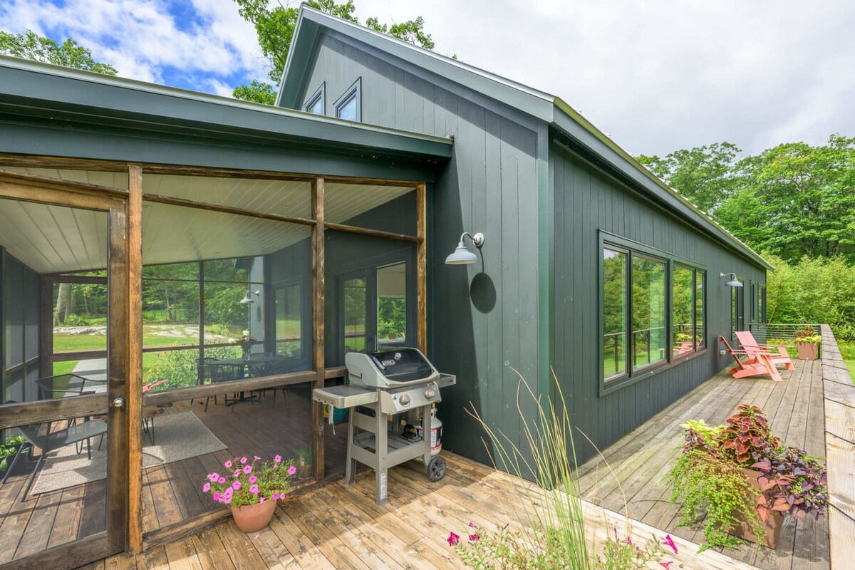 Exterior view of the modern dark green house with a spacious wooden deck. A BBQ grill is positioned on the deck, which also features potted plants and leads to a screened porch.