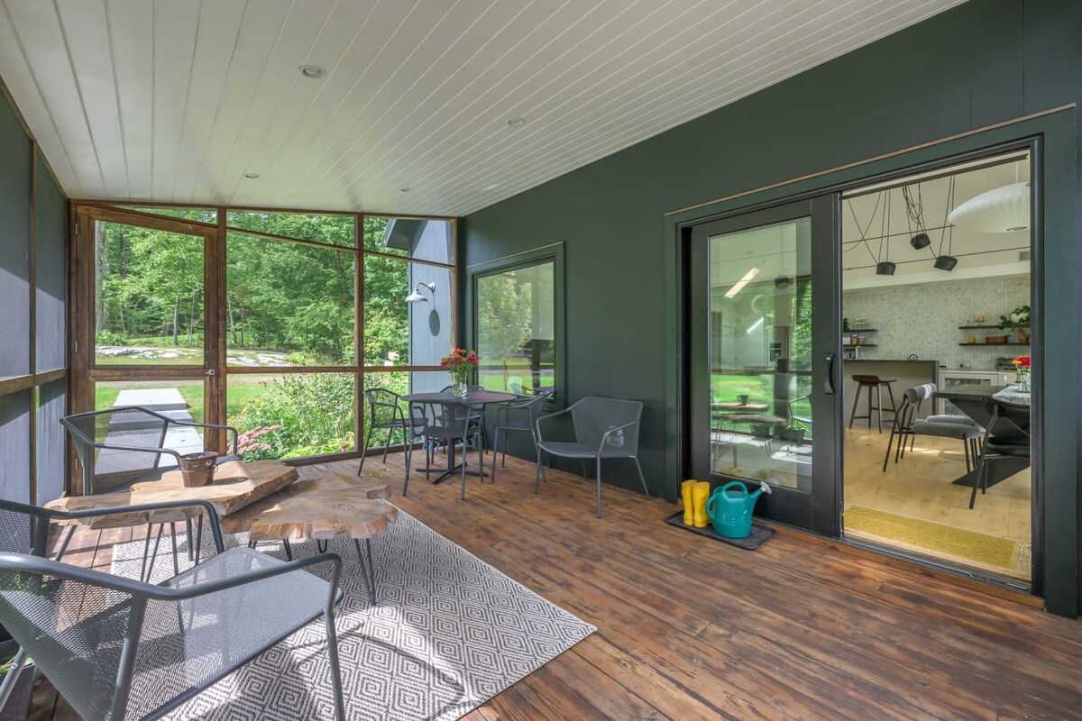 Screened-in porch with dark wood floors and a dark green accent wall, furnished with a small dining table and chairs, and additional lounge seating. Large screens offer views of the lush green outdoors, and sliding glass doors lead into the interior.