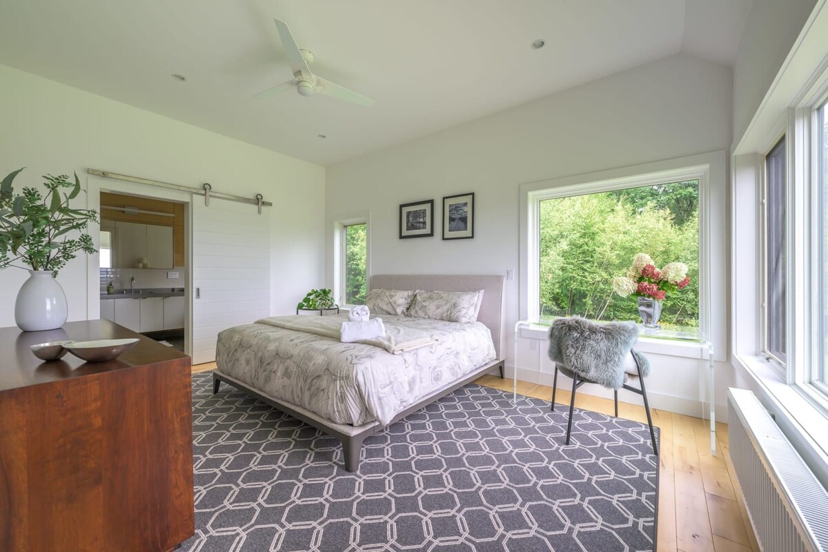 A bright bedroom with light hardwood floors and a vaulted ceiling, featuring a queen-sized bed with patterned bedding, a dark geometric rug, and large windows offering views of lush green trees. A barn door leads to an adjacent room.
