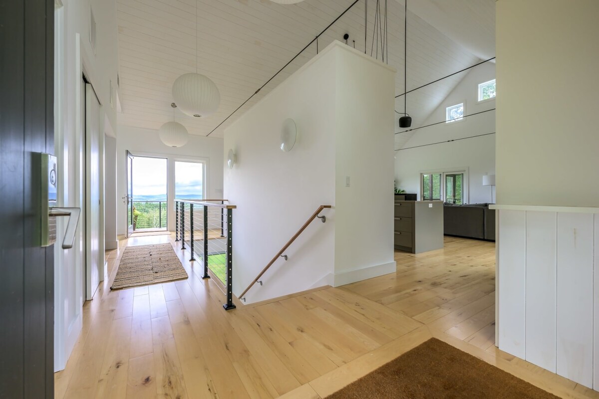 A bright entryway with light hardwood floors and white walls, featuring a modern railing leading downstairs and a large glass door opening to an outdoor balcony with mountain views. Contemporary spherical pendant lights hang from the vaulted ceiling.