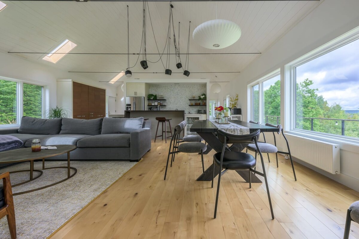 An expansive open-plan living and dining area with light hardwood floors and a vaulted ceiling, featuring a large gray sectional sofa, a dark dining table with chairs, and a modern kitchen bar. Large windows provide abundant natural light and views of the surrounding landscape.