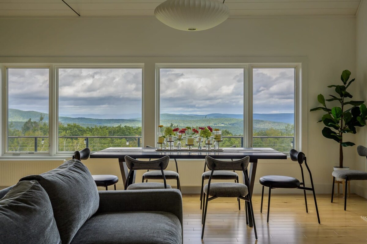 Elegant dining area with a long dark dining table and six modern chairs, positioned in front of large windows that offer stunning panoramic views of rolling green hills and distant mountains under a cloudy sky.