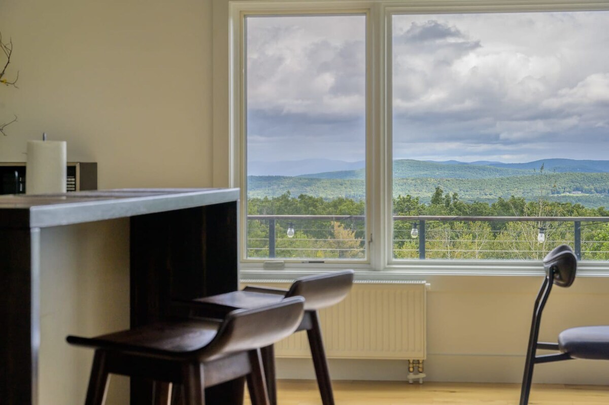 A close-up view of a kitchen bar area with dark wooden stools, positioned in front of large windows that offer expansive views of distant mountains and a verdant landscape under a cloudy sky. A white radiator is visible below the windows.