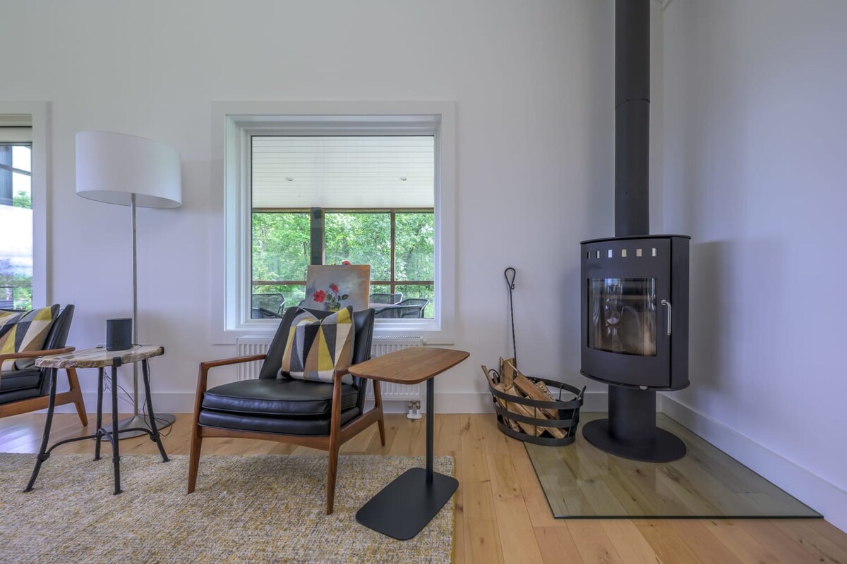 A cozy corner of a modern living room features a black leather armchair with a geometric pillow, a floor lamp, and a black wood-burning stove on a glass hearth, all set against white walls and light hardwood floors. A large window offers a glimpse of lush greenery outside.