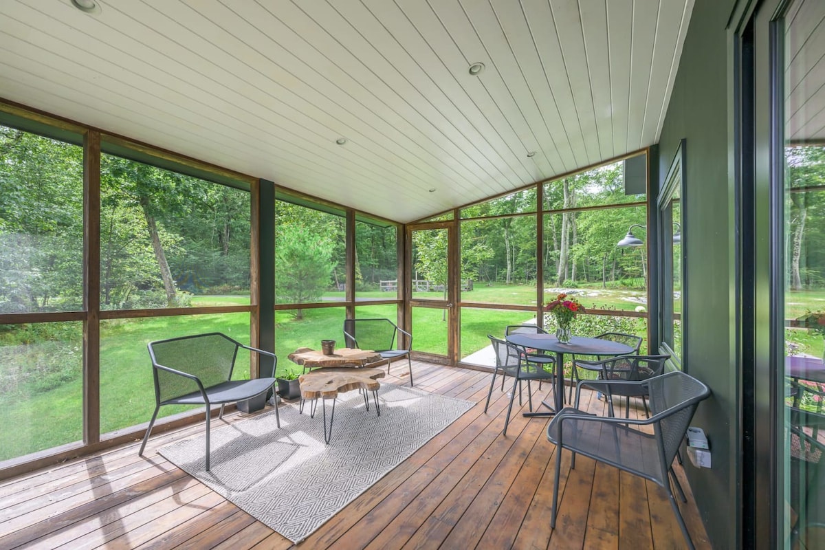 Spacious screened porch with a wooden floor, furnished with two dark metal armchairs and a small wooden coffee table on a rug, and a dining set with a round table and chairs, all surrounded by screens offering views of a lush green lawn and trees.