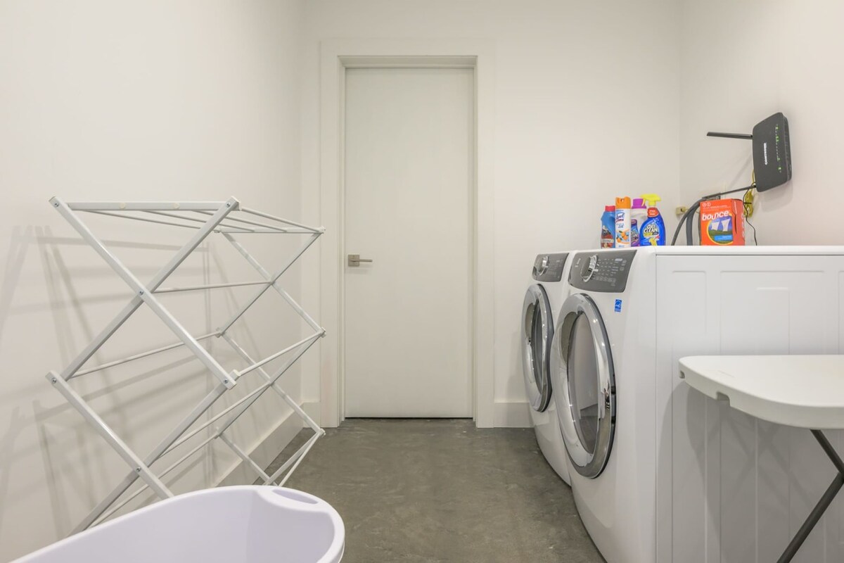 Bright laundry room with white walls and a concrete floor, featuring a white front-loading washer and dryer set. A white drying rack stands nearby, and laundry supplies are neatly arranged on top of the machines.