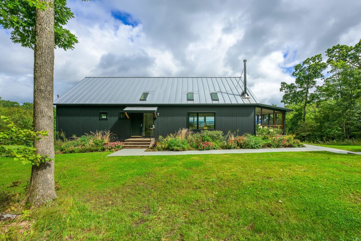 The front exterior of a modern barn-style house features dark siding, a dark metal roof with skylights, and a prominent chimney. Lush garden beds with various plants line the walkway to the entrance, set against a vibrant green lawn and mature trees.
