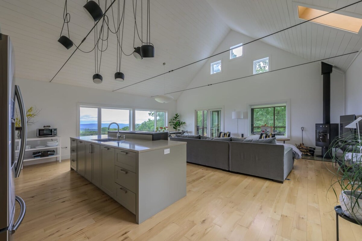 A spacious, open-concept living area with a high vaulted ceiling and light hardwood floors, featuring a large gray sectional sofa, a modern kitchen island, and a black wood-burning stove. Large windows offer panoramic views of mountains and lush trees.