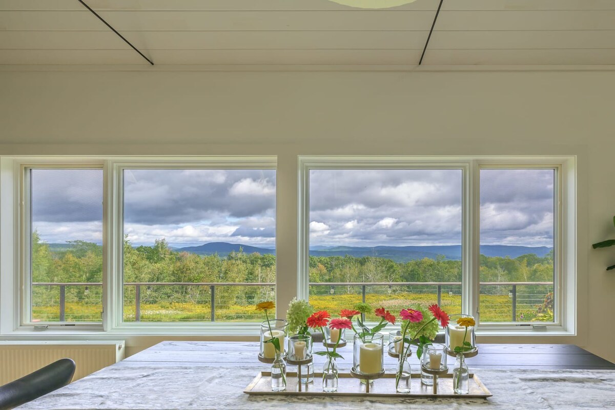 A close-up of a dining table adorned with a floral centerpiece, positioned in front of expansive windows that offer breathtaking panoramic views of rolling green mountains and a cloudy sky. The room features light hardwood floors and white walls.
