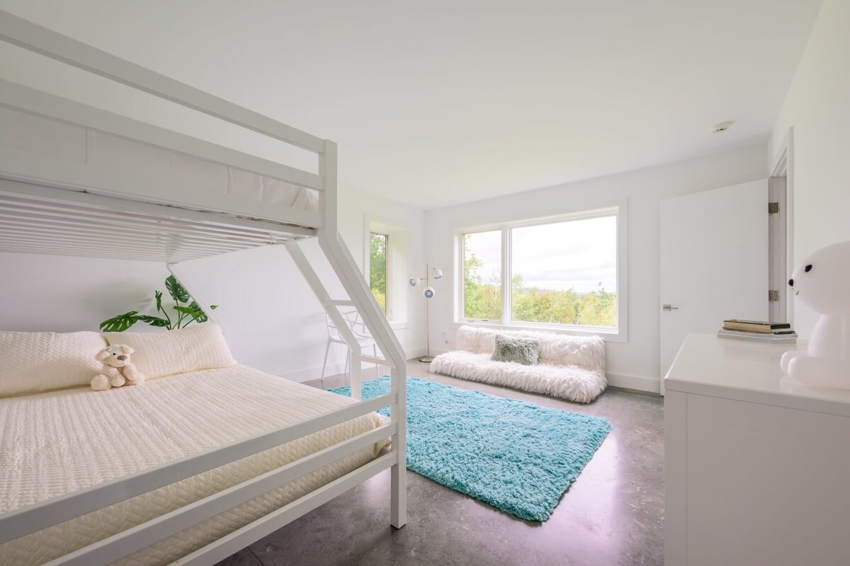 Spacious bedroom with a white metal bunk bed, polished concrete floors, a vibrant turquoise area rug, and a window offering views of the outdoors.