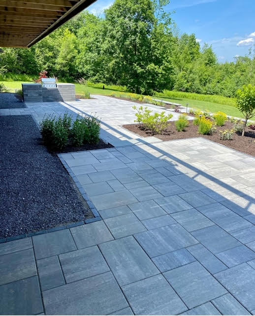 Expansive stone paver patio with a dark gravel border, extending from beneath the house. Landscaped garden beds with small shrubs line the patio, leading towards a distant green landscape.