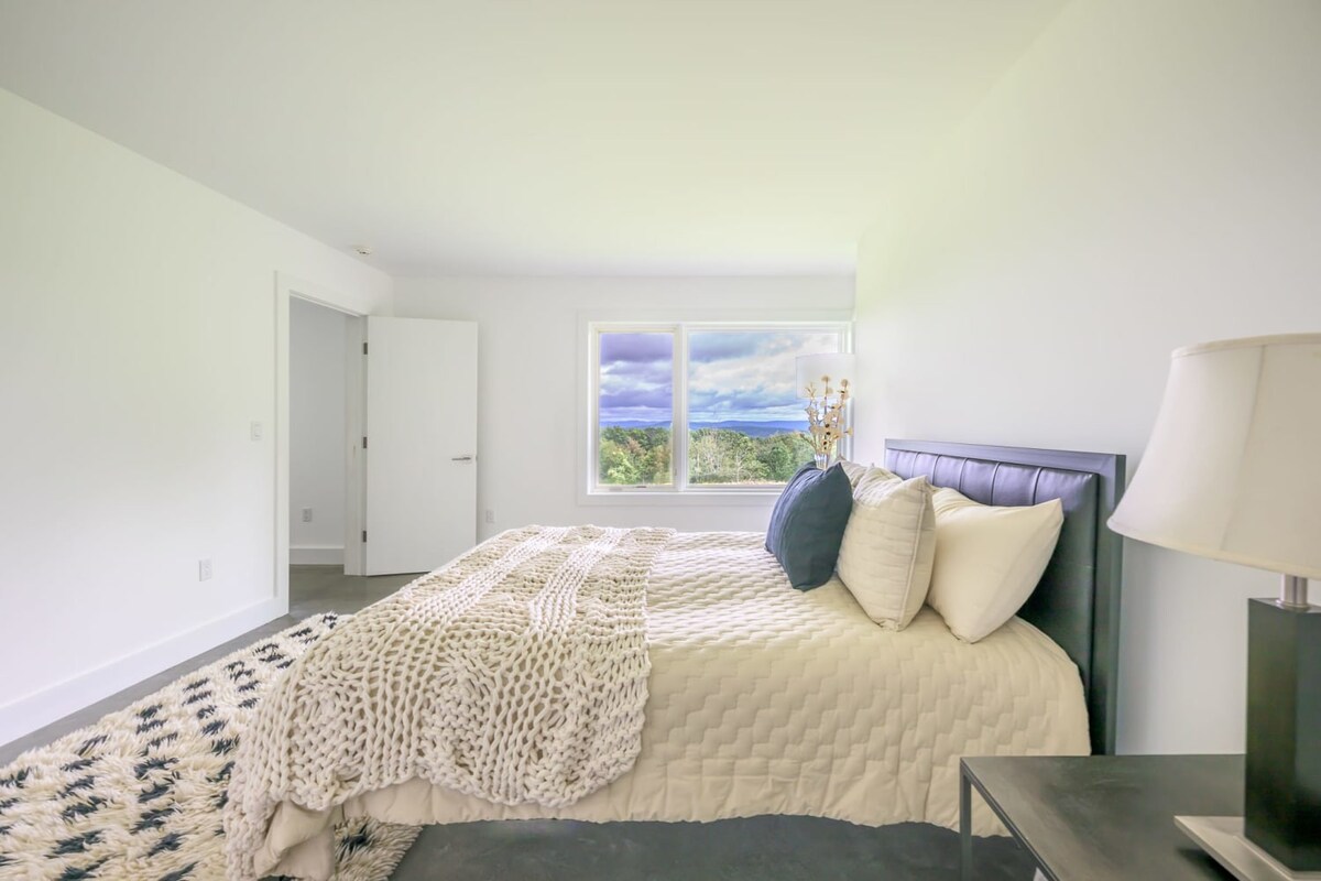 Cozy bedroom featuring a black bed frame with neutral bedding, polished concrete floors, a black and white patterned rug, and a large window with a scenic view.