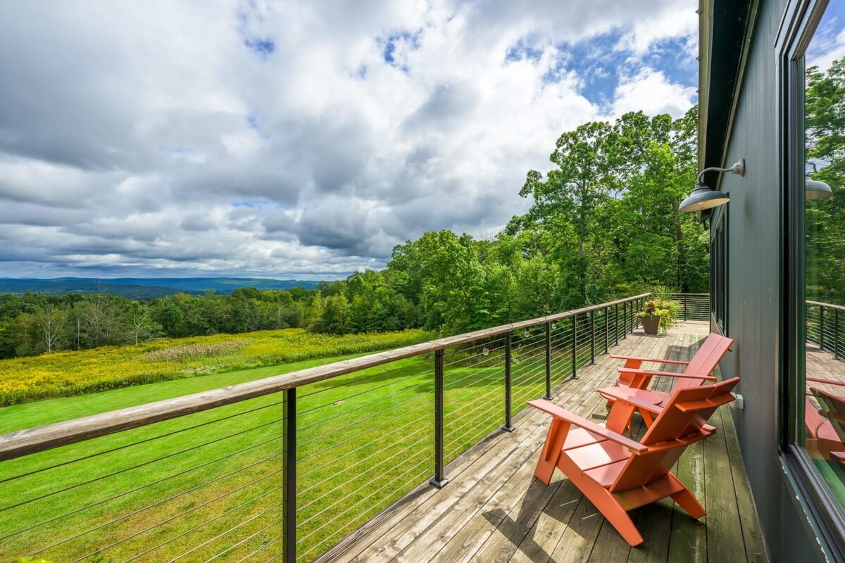 Expansive wooden deck with cable railings, offering panoramic views of rolling green hills and distant mountains under a cloudy sky. Two orange Adirondack chairs invite relaxation, facing the scenic landscape.