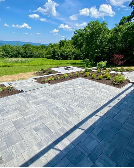 Wide view of the multi-level stone paver patio and landscaped garden beds, with a square fire pit visible in the distance. The patio offers open views of a vast green field and distant mountains under a partly cloudy sky.