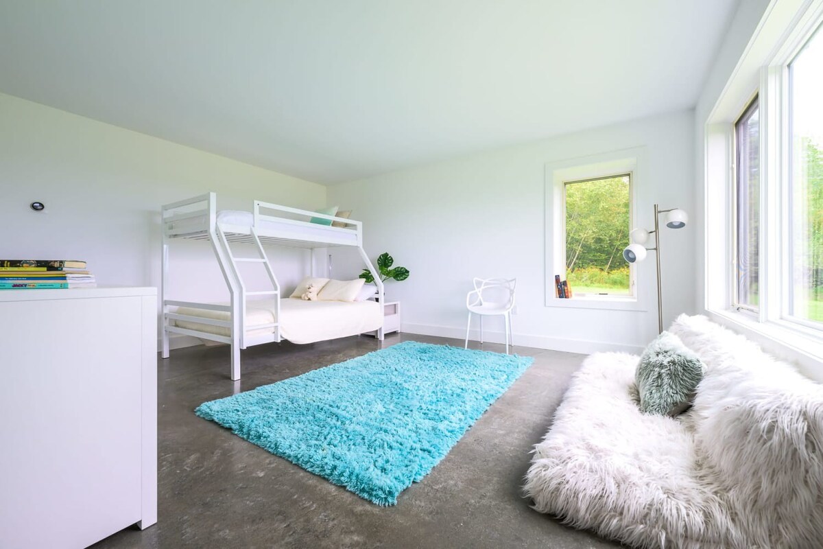 Bright bedroom with polished concrete floors, a white metal bunk bed, a fluffy turquoise area rug, and a cozy white faux fur floor cushion by a large window.