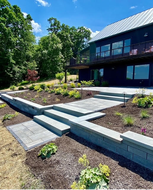 Landscaped backyard with a multi-level stone paver patio and retaining walls, leading up to the dark-colored modern house. Lush greenery and steps are integrated into the design, creating an inviting outdoor space.