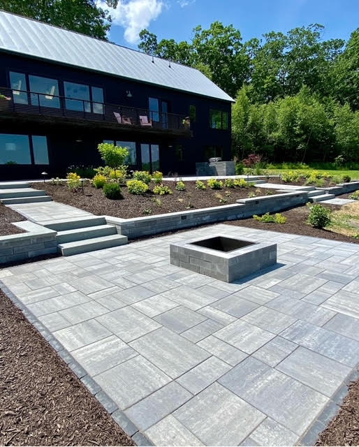 Spacious stone paver patio featuring a built-in square fire pit, surrounded by landscaped garden beds and retaining walls. The modern dark house is visible in the background, with lush trees under a bright sky.