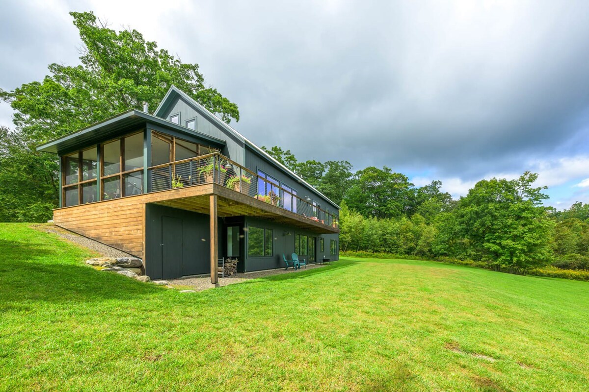This view of the modern dark-sided house showcases an elevated wooden deck and a screened-in porch on the upper level, overlooking a vast, sloping green lawn. The lower level features additional windows and an entrance, with dense foliage and trees surrounding the property.