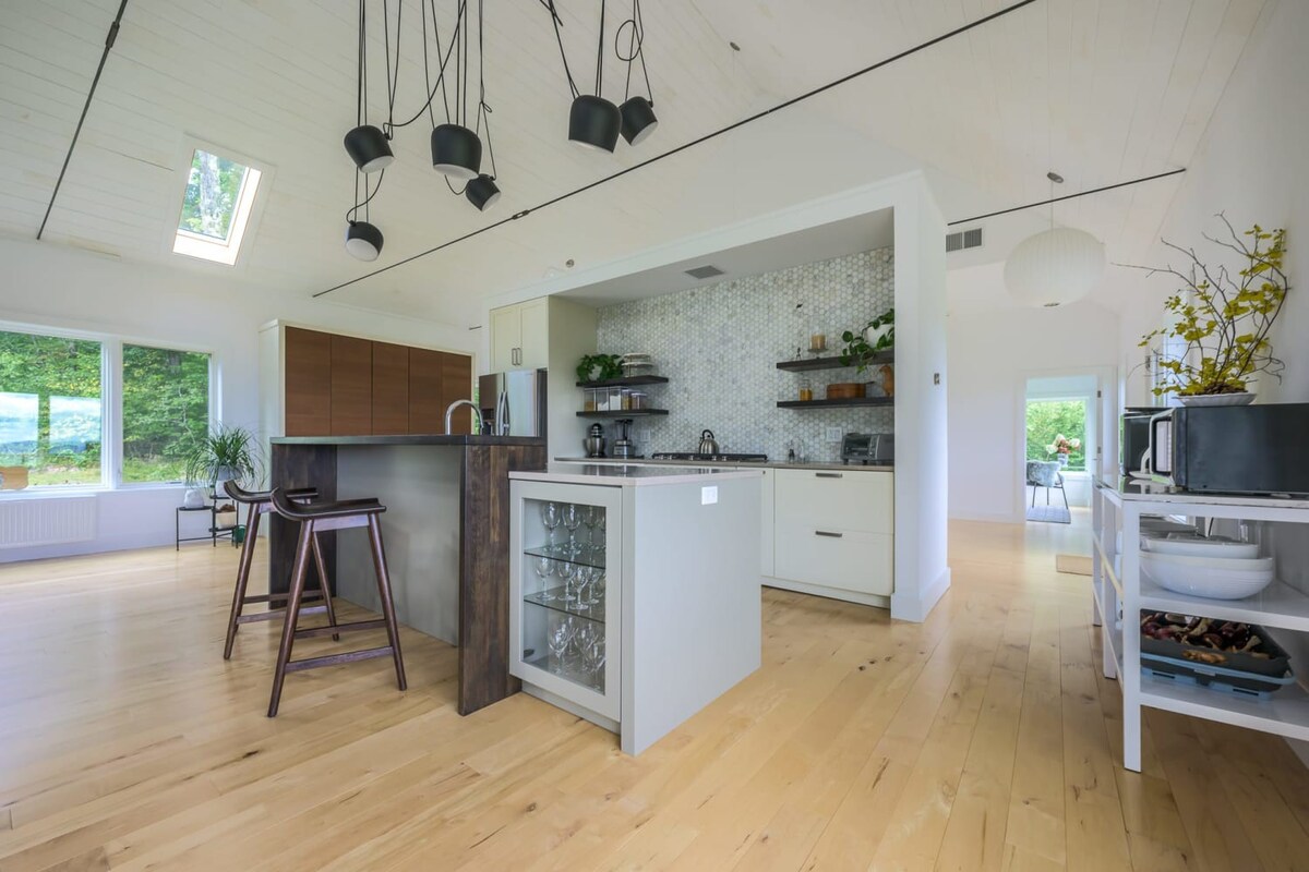A modern kitchen with light hardwood floors and a vaulted ceiling, featuring a large island with bar seating, white cabinetry, a tiled backsplash, and open shelving. Large windows overlook lush greenery, and unique black pendant lights hang overhead.