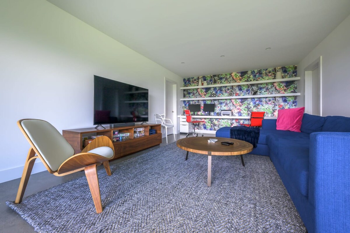 Modern living room with a large blue sectional sofa, a wooden coffee table, and a mid-century modern chair. A large TV is mounted above a media console, and a vibrant floral wallpaper accentuates a built-in desk area with two red office chairs.