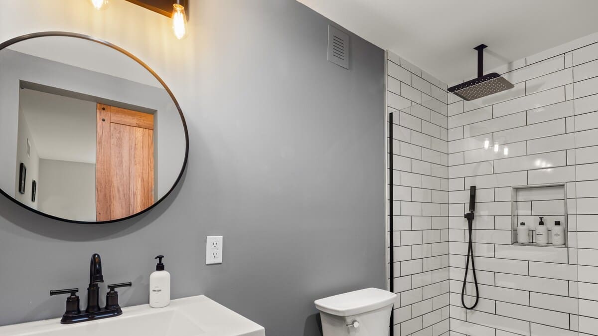 Modern bathroom with a white subway tile shower, black rain shower head and handheld sprayer, a toilet, and a dark gray vanity with a round mirror.