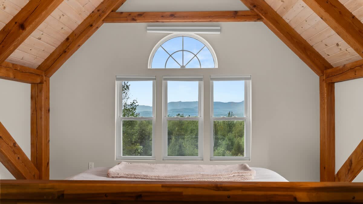A close-up view from the bedroom highlights the stunning mountain landscape visible through a large triple window set, topped by an elegant arched transom. The room features a vaulted ceiling with exposed wooden beams.