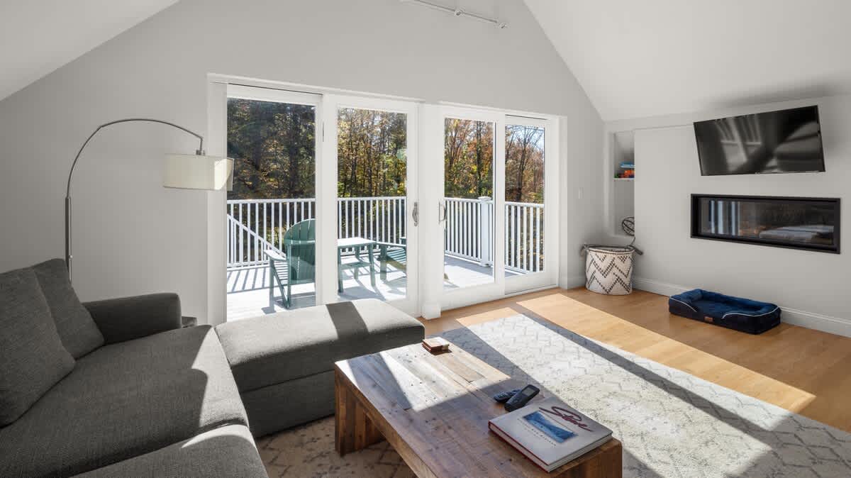 Bright living room with vaulted ceilings, light hardwood floors, a large gray sectional sofa, and a rustic wooden coffee table. Sliding glass doors open to a balcony with outdoor seating, offering views of surrounding trees.