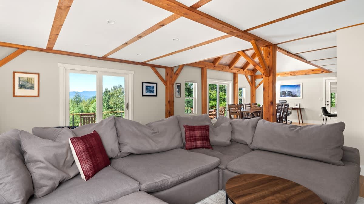 A cozy living room featuring a large gray sectional sofa with red plaid throw pillows, light hardwood floors, and exposed wooden beams on the ceiling, with sliding glass doors offering mountain views.
