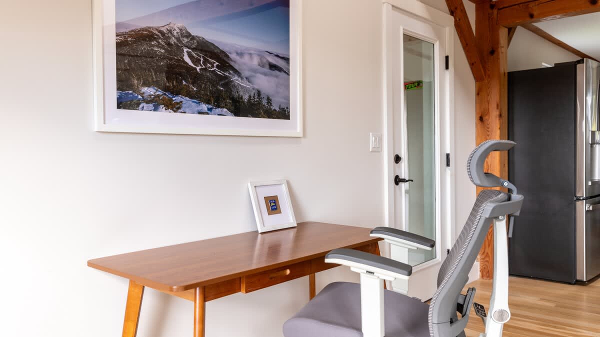 A dedicated office nook features a sleek wooden desk and an ergonomic grey office chair, positioned against a light wall adorned with a large framed mountain landscape print. A glass-paned door is visible to the right.