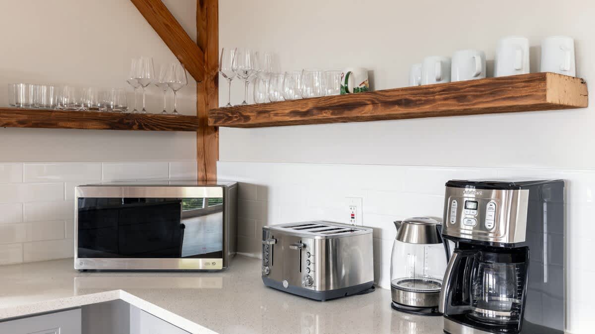 A section of the kitchen counter showcasing a stainless steel microwave, a toaster, a coffee maker, and an electric kettle, all neatly arranged beneath open wooden shelving with glassware.
