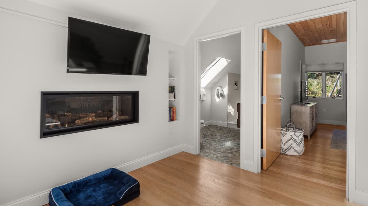 Modern living room with light hardwood floors, a wall-mounted flat-screen TV above a linear gas fireplace, and a cozy dog bed. A doorway leads to an adjacent room with a skylight and tiled floor.